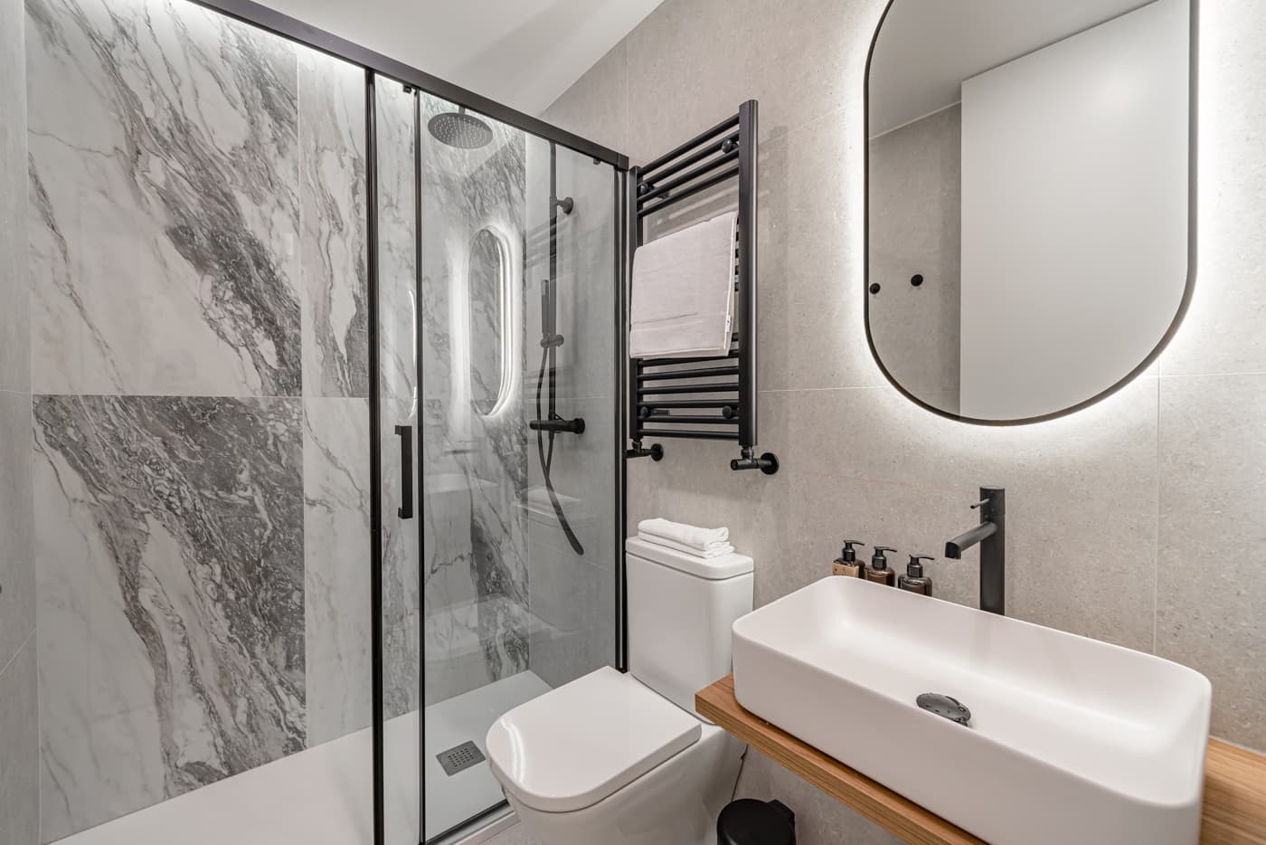 Baño con ducha de mampara, revestimiento gris veteado y espejo retroiluminado. Atocha 94, Madrid.