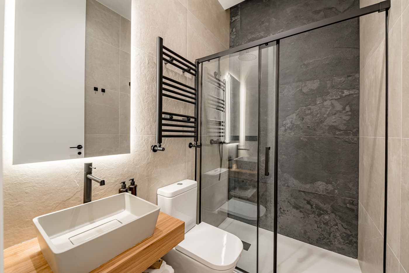 Baño con ducha de mampara negra, revestimiento oscuro y lavabo sobre encimera de madera. Divino Pastor 14, Madrid.