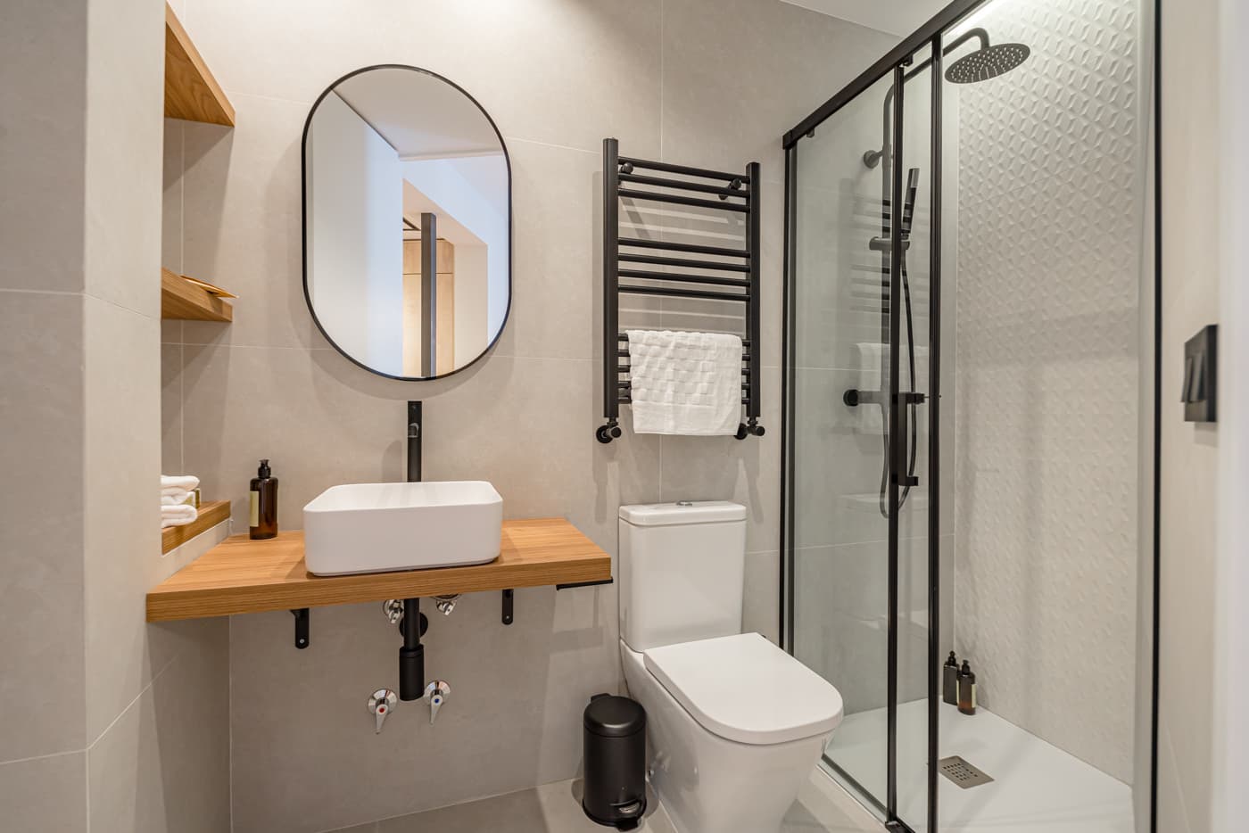 Baño con ducha de mampara negra, espejo ovalado y lavabo sobre encimera de madera. Hermosilla 133, Madrid.