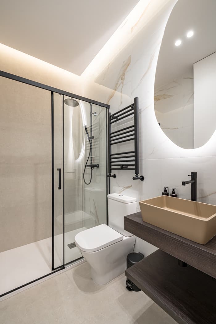 Baño con ducha de mampara, espejo retroiluminado y lavabo sobre encimera oscura. Jesús del Valle 25, Madrid.