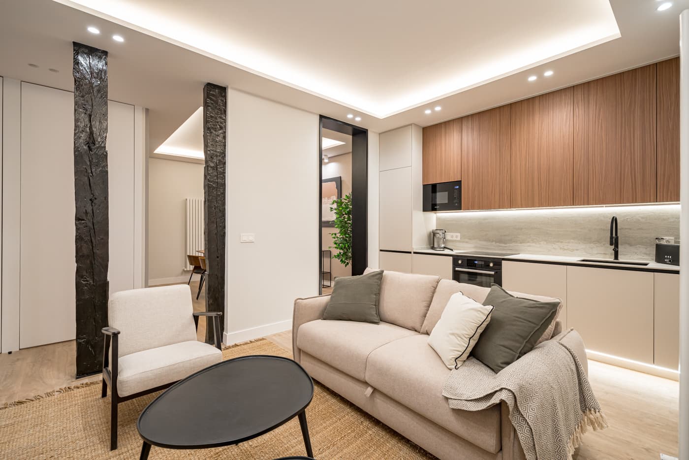 Salón abierto a la cocina, con muebles altos en acabado madera y dos pies derechos de madera antigua a la vista. Jesús del Valle 25, Madrid.