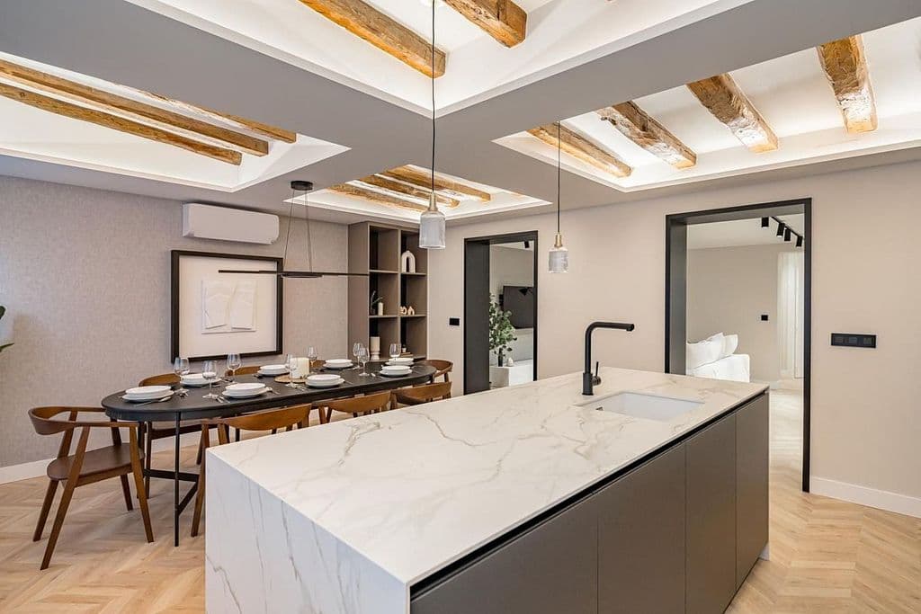 Isla de cocina con encimera clara veteada en primer plano; al fondo, el comedor bajo las vigas vistas. Mayor 73, Madrid.