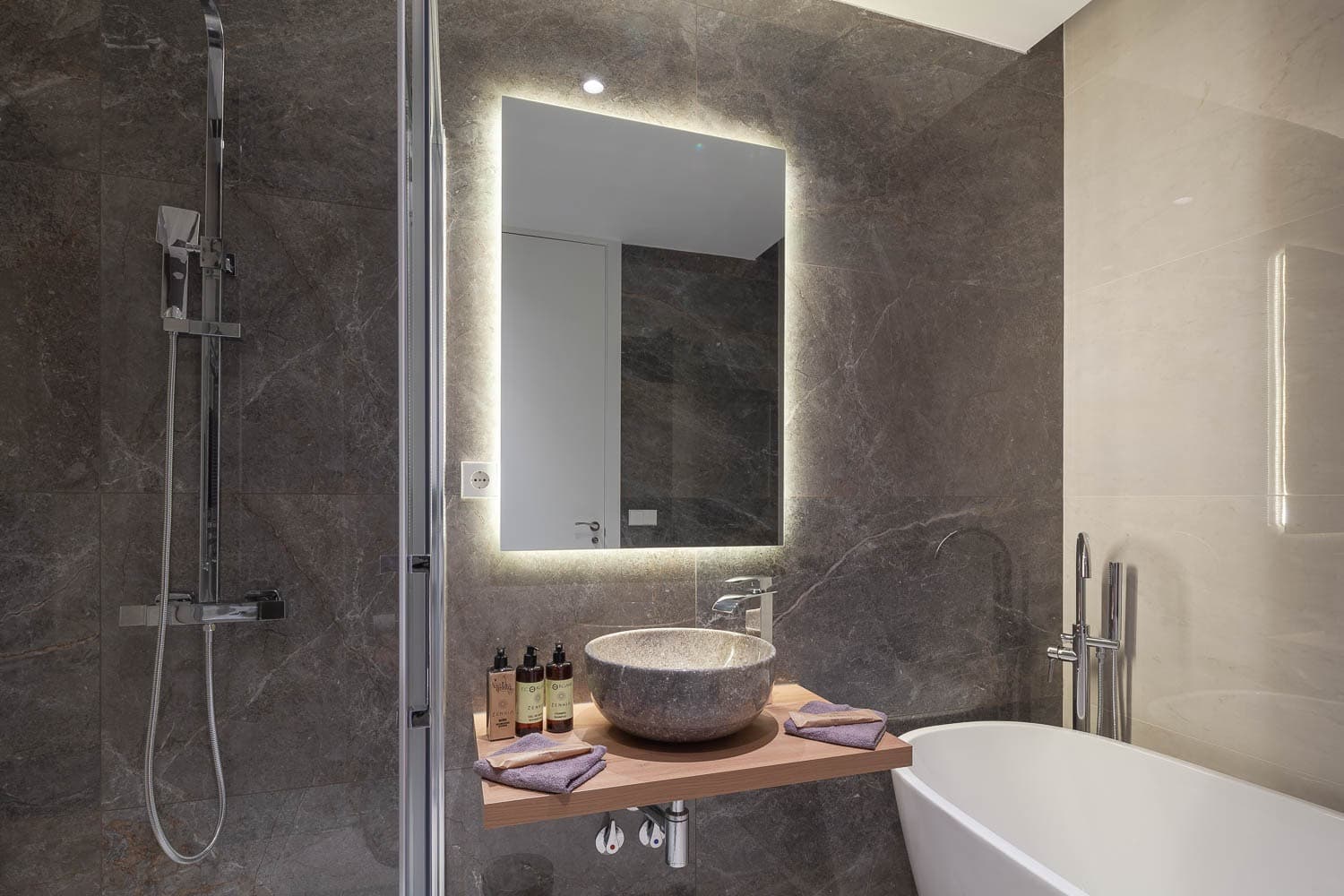 Baño con revestimiento gris veteado, espejo retroiluminado, lavabo sobre encimera y bañera exenta. Calle Ruiz, Madrid.