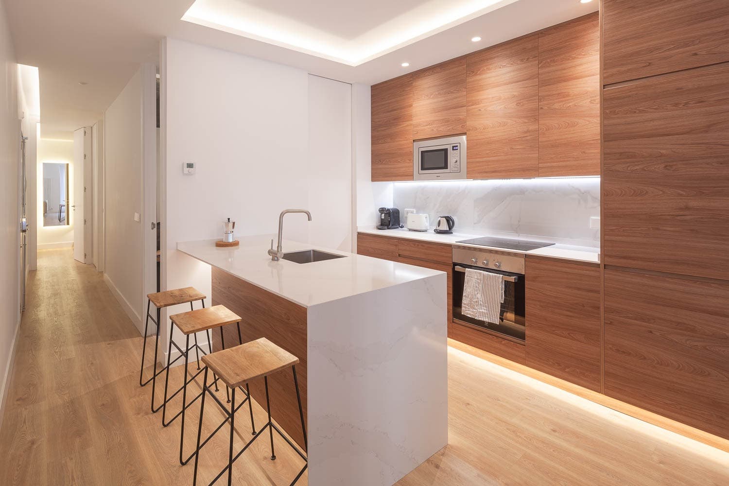 Cocina con península, frentes en acabado madera y pasillo al fondo. Calle Ruiz, Madrid.