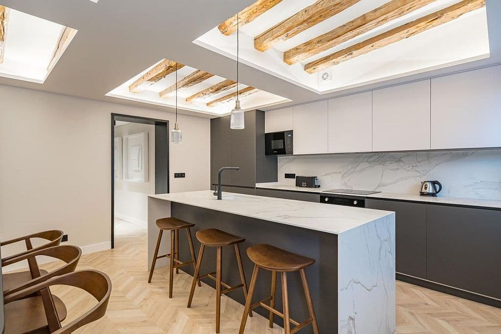 Cocina abierta con isla y taburetes; en el techo, vigas de madera vistas dentro de dos foseados iluminados. Mayor 73, Madrid.