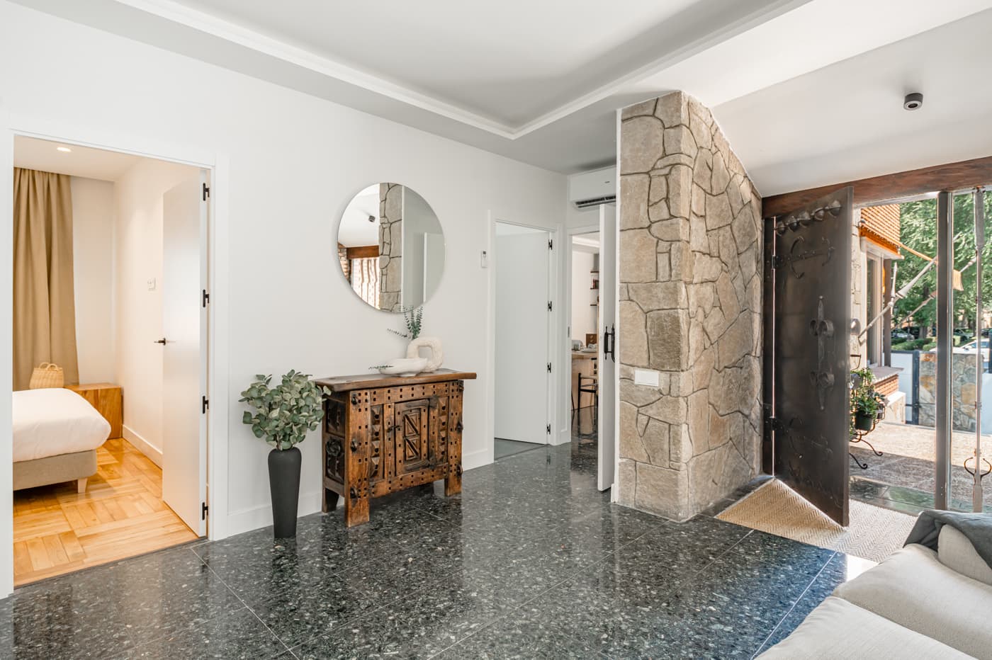 Recibidor con muro de piedra y puerta de entrada con herrajes de forja; a la izquierda, un dormitorio. Vía Complutense, Alcalá de Henares.