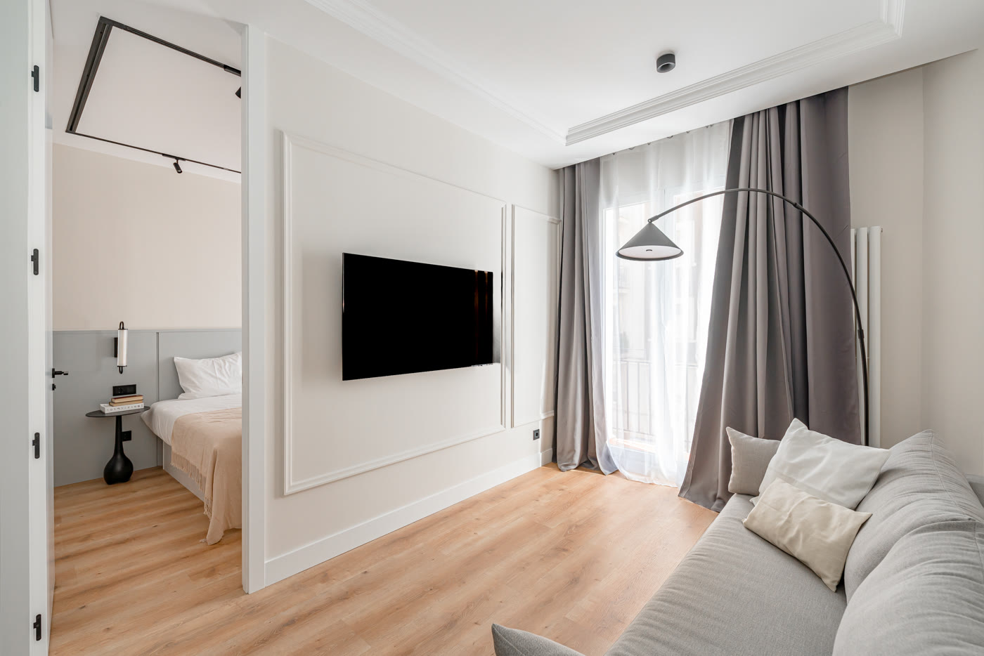 Salón con sofá gris y lámpara de arco, con el dormitorio visible al fondo